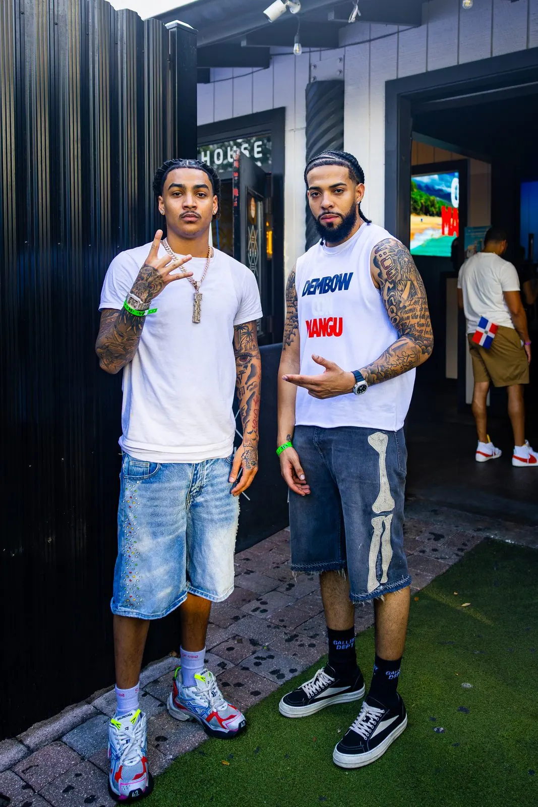 Two men standing on the paved and turfed strip outside an open doorway, green entry wristbands on both their wrists, a black corrugated wall beside them and a lit room behind.