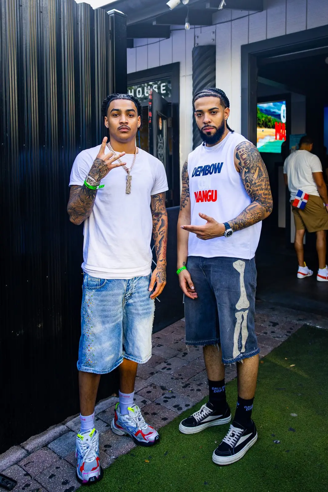 Two men standing side by side on a strip of turf, one in a white vest printed DEMBOW CON MANGÚ.