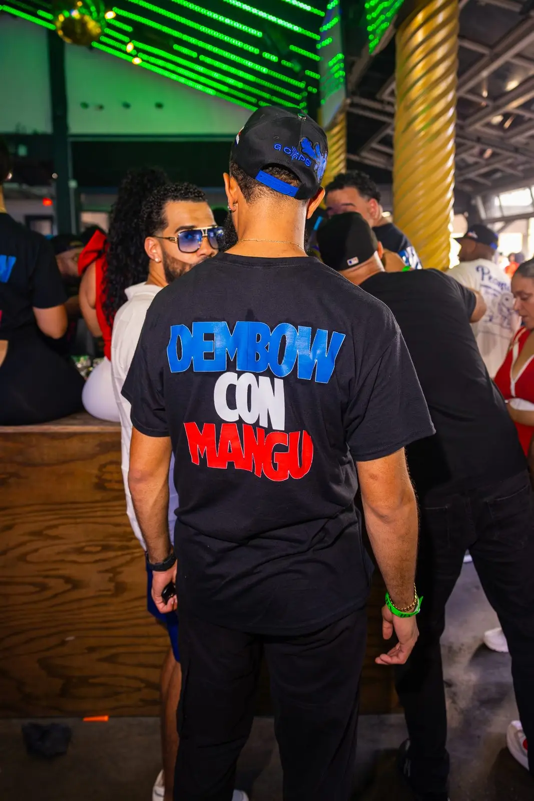The back of a black DEMBOW CON MANGÚ T-shirt in the middle of a crowd, green neon running the length of the ceiling.