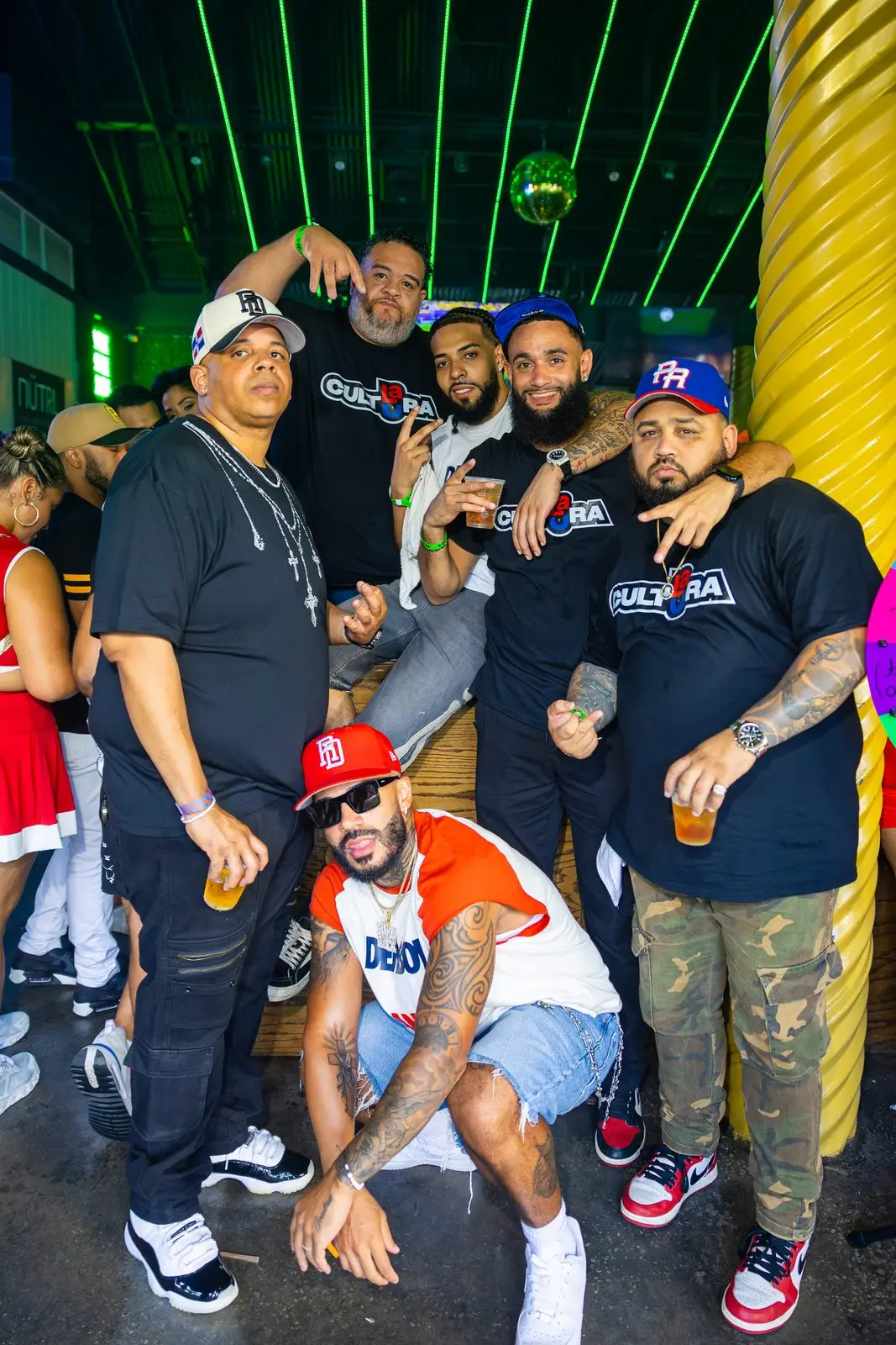 A group of friends packed together for a photograph under a green-lit ceiling, drinks in hand, several in matching CULTURA shirts.