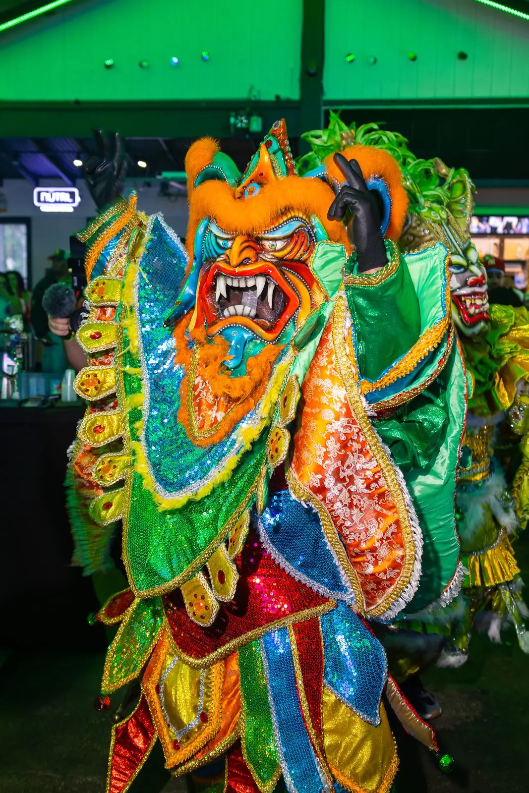 Carnival costumes at close range: horned devil masks with bared teeth, over capes of orange, green and gold sequins.