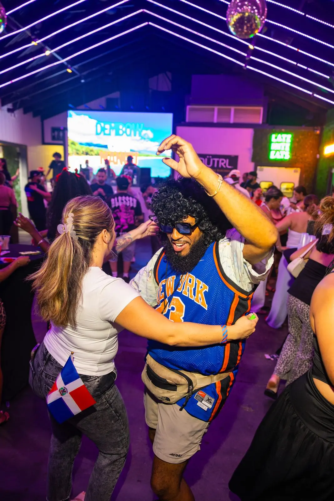 A couple dancing together in the middle of the floor under strips of white ceiling neon, a small Dominican flag in the back pocket of the woman beside them.