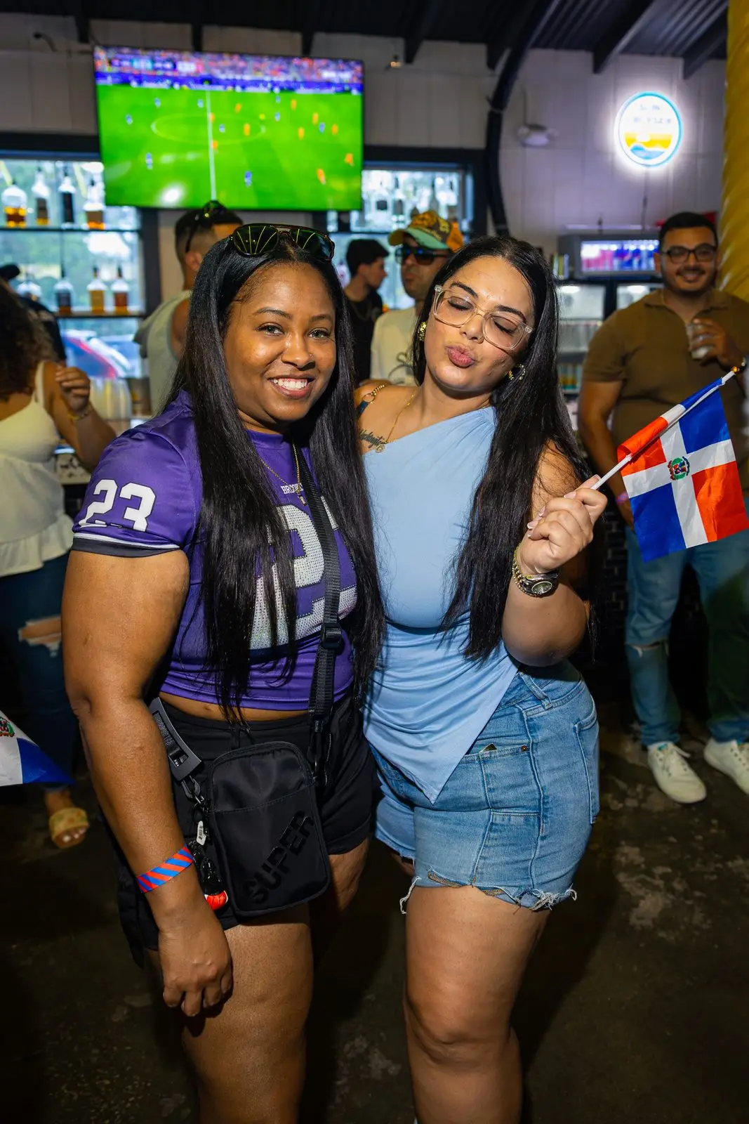 Two women standing together for a photograph, one in a purple jersey and one holding a small Dominican flag up beside them, a football match on the screen behind.