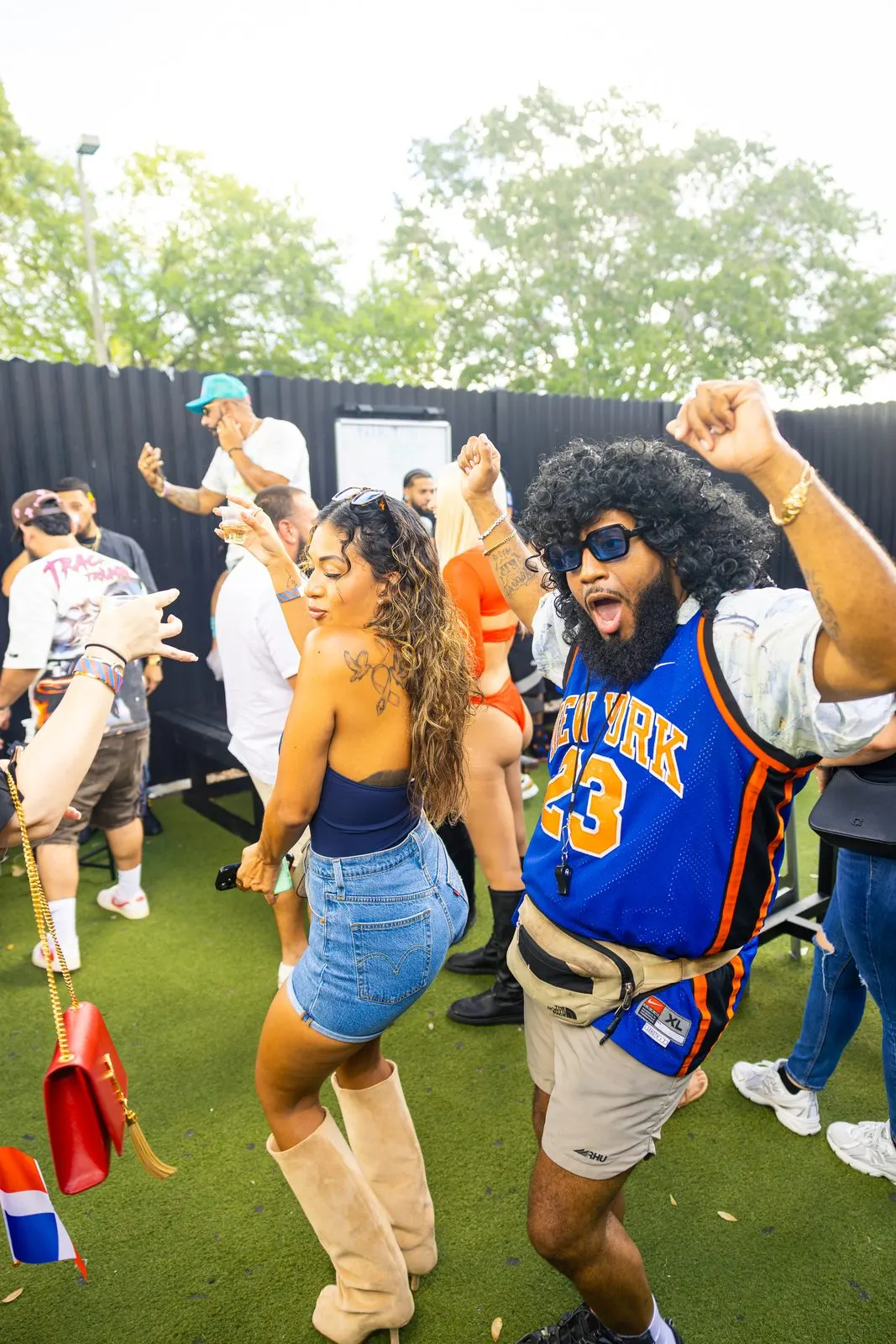 Two people dancing outdoors on green turf in daylight, arms up, trees and a dark fence line behind them.