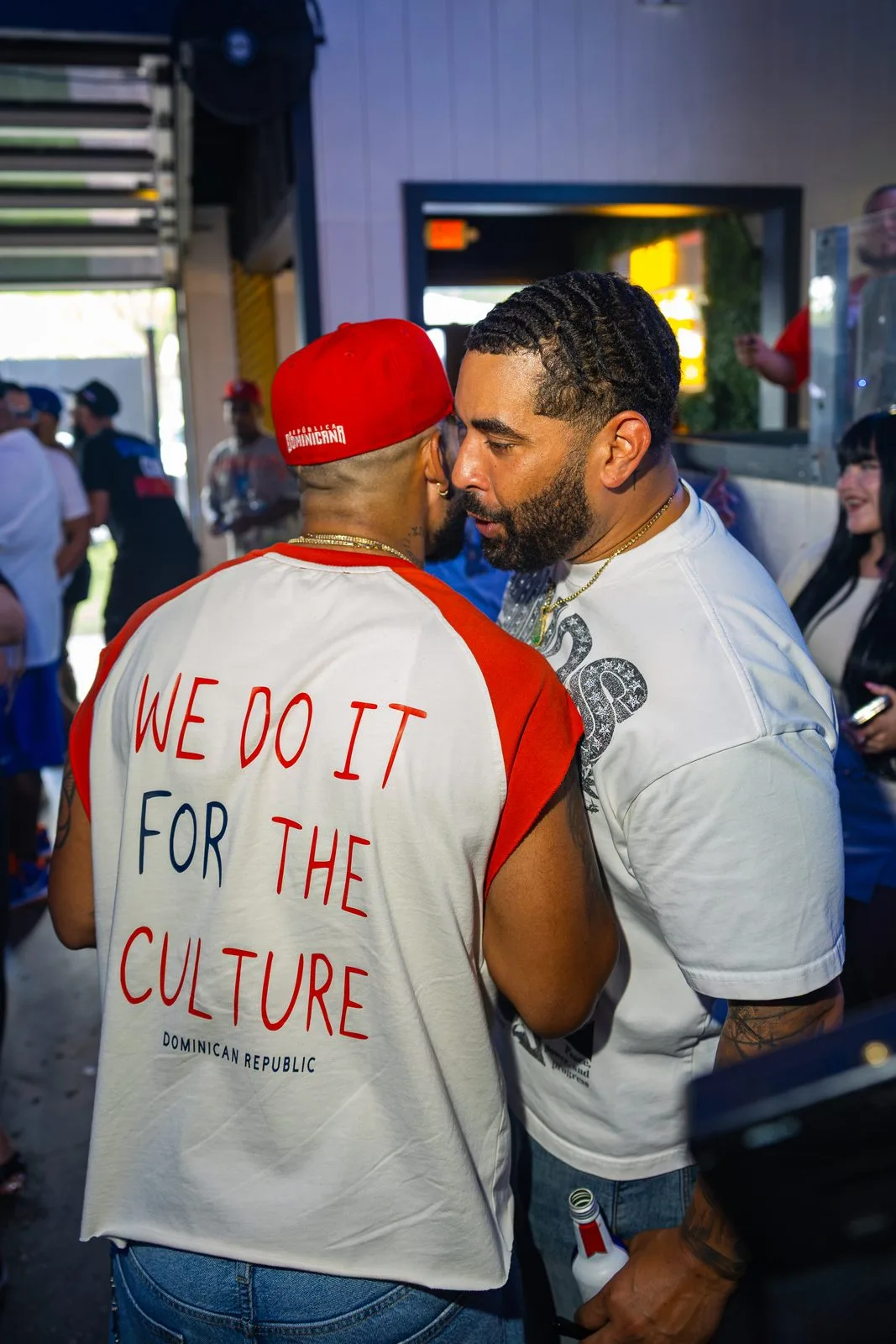 Two men leaning in to greet each other in a packed room, one in a red cap, the other's shirt printed WE DO IT FOR THE CULTURE.