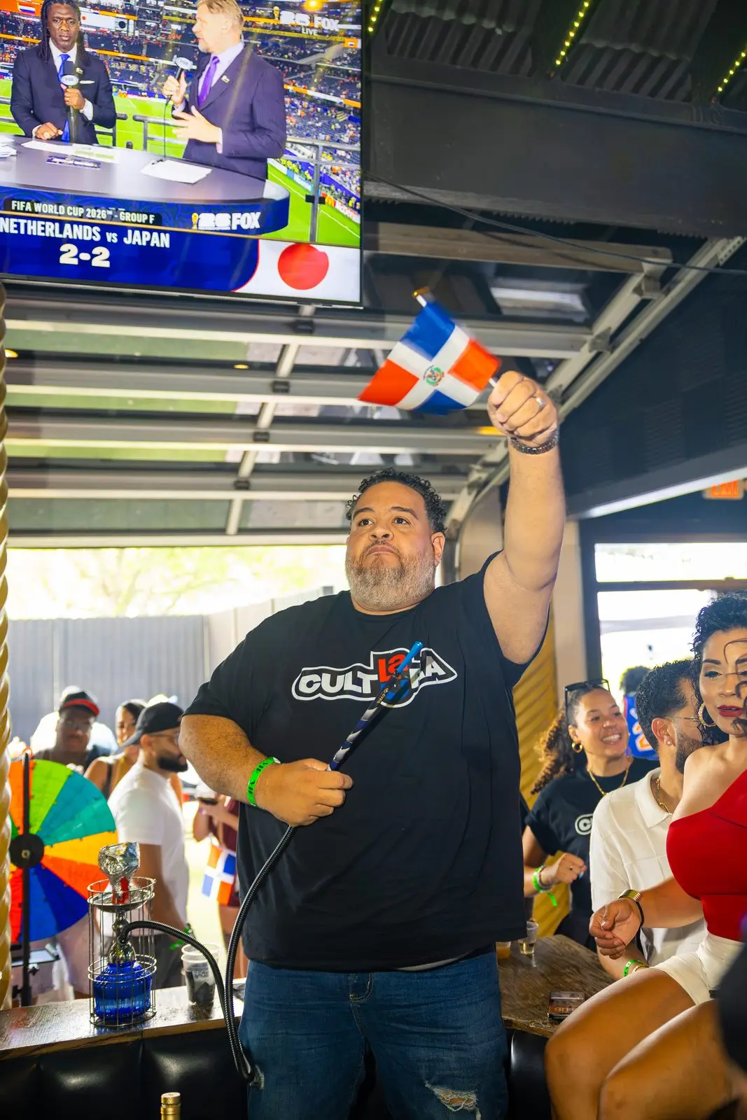 A man in a CULTURA shirt with one arm raised, holding a small Dominican flag above the crowd.