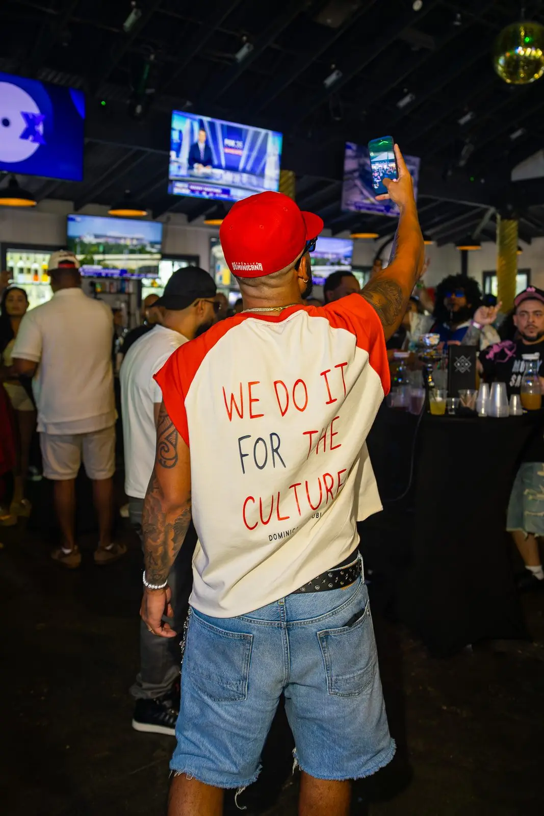 A man seen from behind in a red cap with his phone held up to film, the back of his shirt reading WE DO IT FOR THE CULTURE.