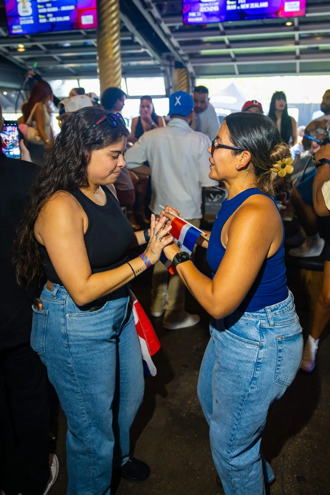 Two women standing close together in the crowd, passing a small Dominican flag between their hands.