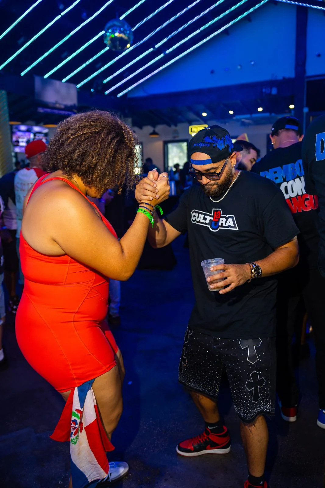 Two people dancing hand in hand on a floor lit blue, a Dominican flag knotted at her waist, Dembow Con Mangú shirts in the crowd behind them.