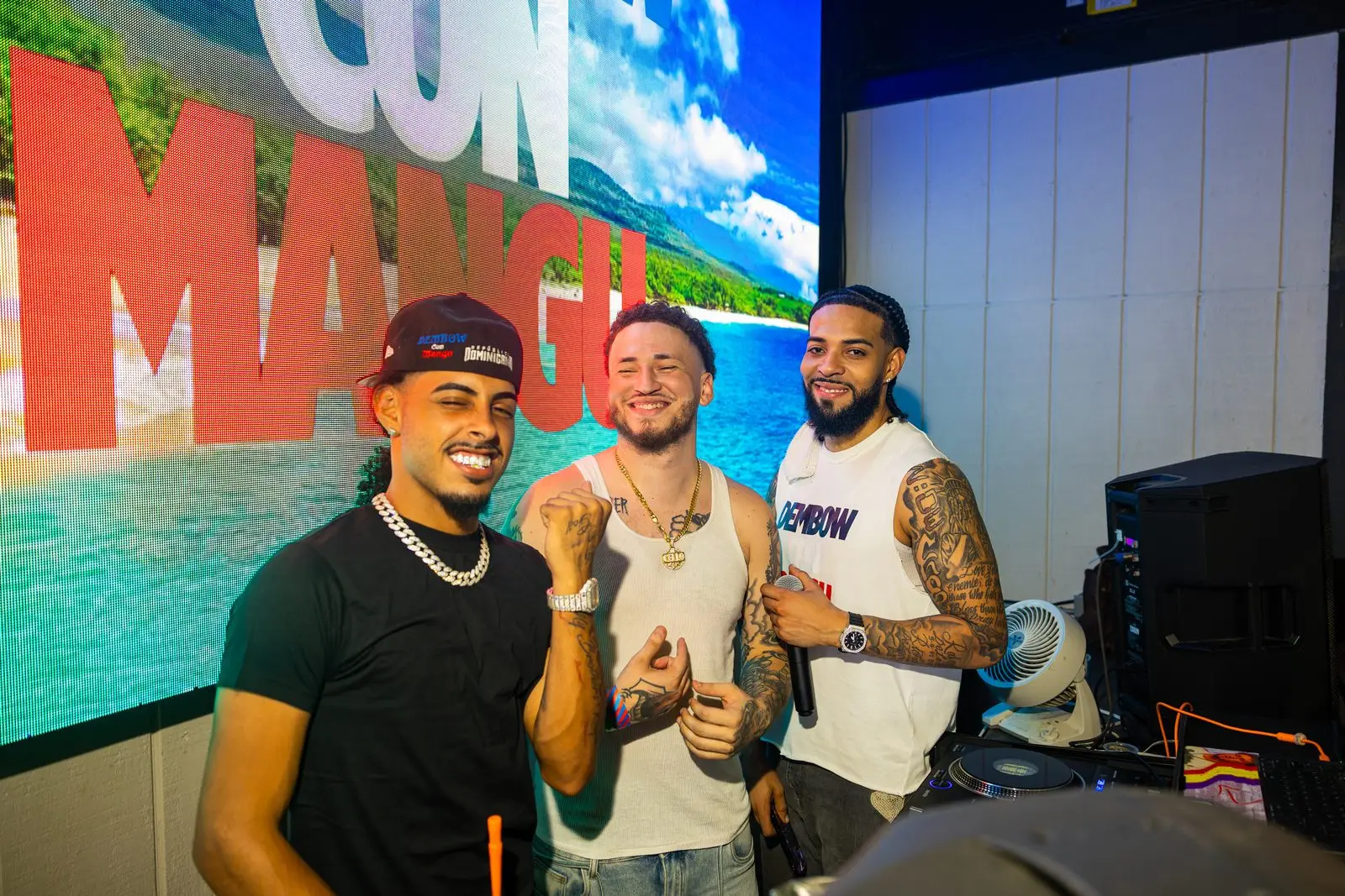 Three men standing shoulder to shoulder in front of a wall-sized screen showing a beach and the words CON MANGÚ in red block letters.