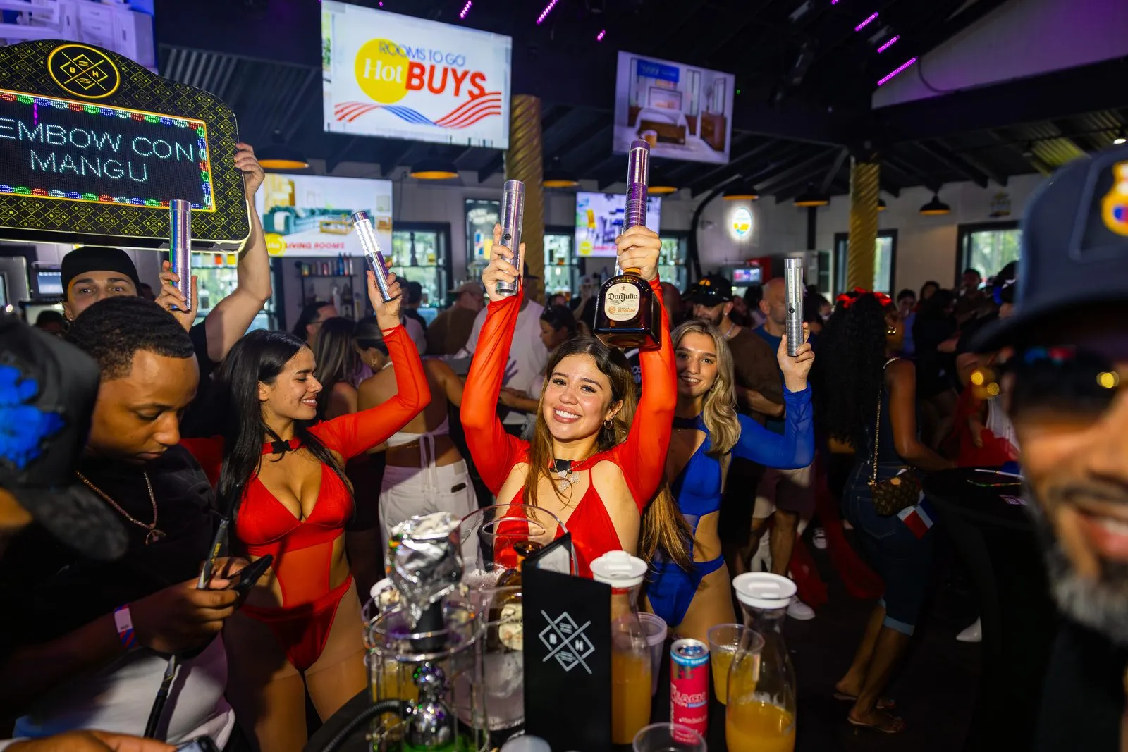 A bottle held up over an ice bucket at a Dembow Con Mangú table, cups and drinks on the table, the group around it with their hands in the air.