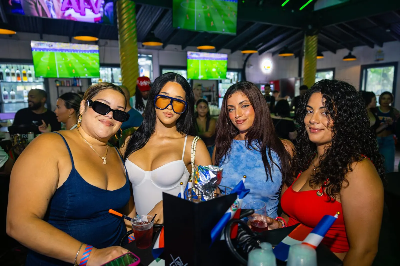Four women posed together at the bar with drinks, small Dominican flags on the counter in front of them and screens on the wall behind.