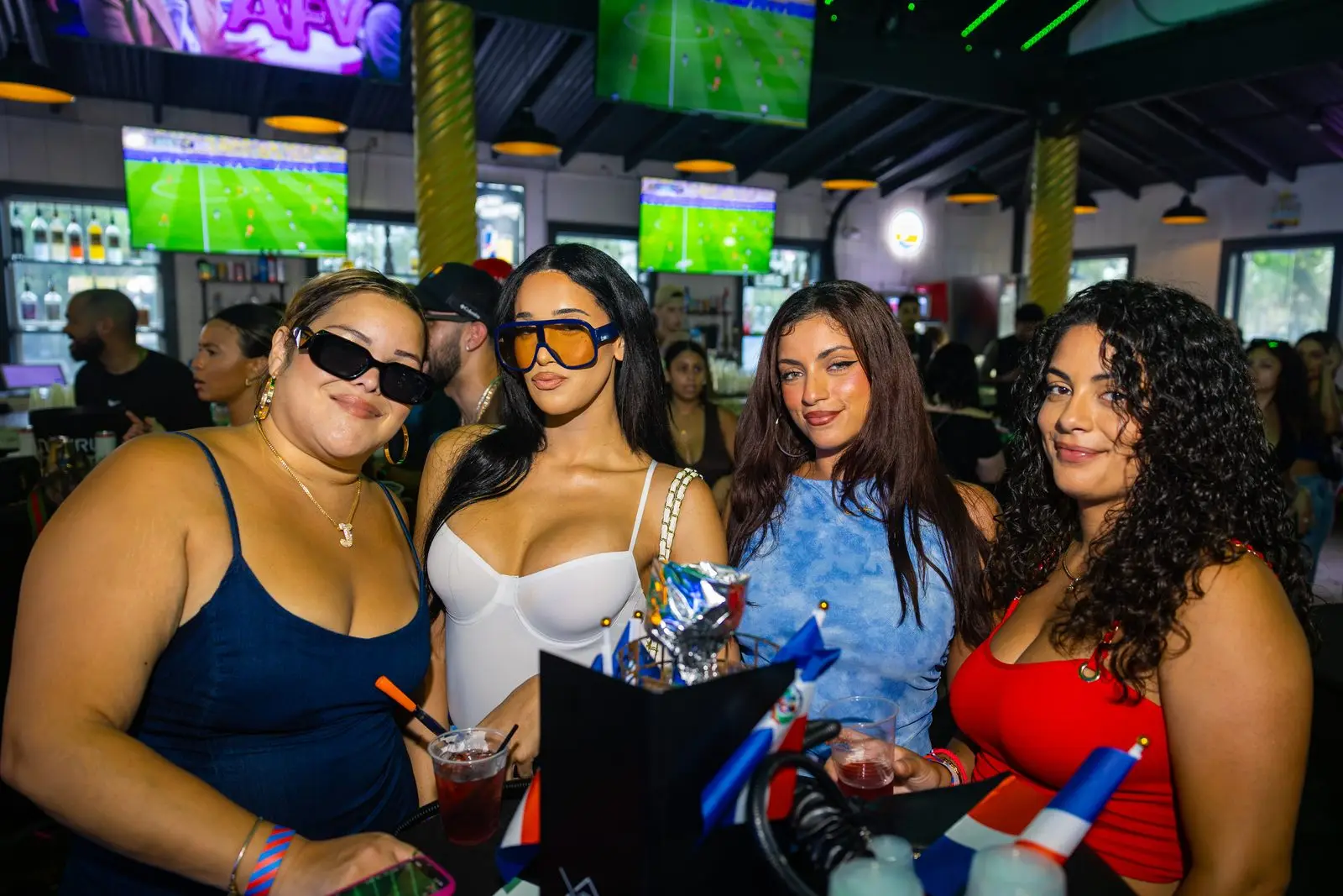 Four women posed shoulder to shoulder at the bar with drinks in their hands, one of them holding a small Dominican flag, televisions and daylight through the shutters behind them.