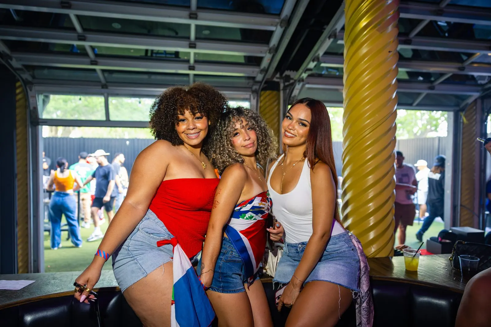 Three friends posed together at the bar rail in red, white and blue, an open roll-up door onto grass and daylight behind them.