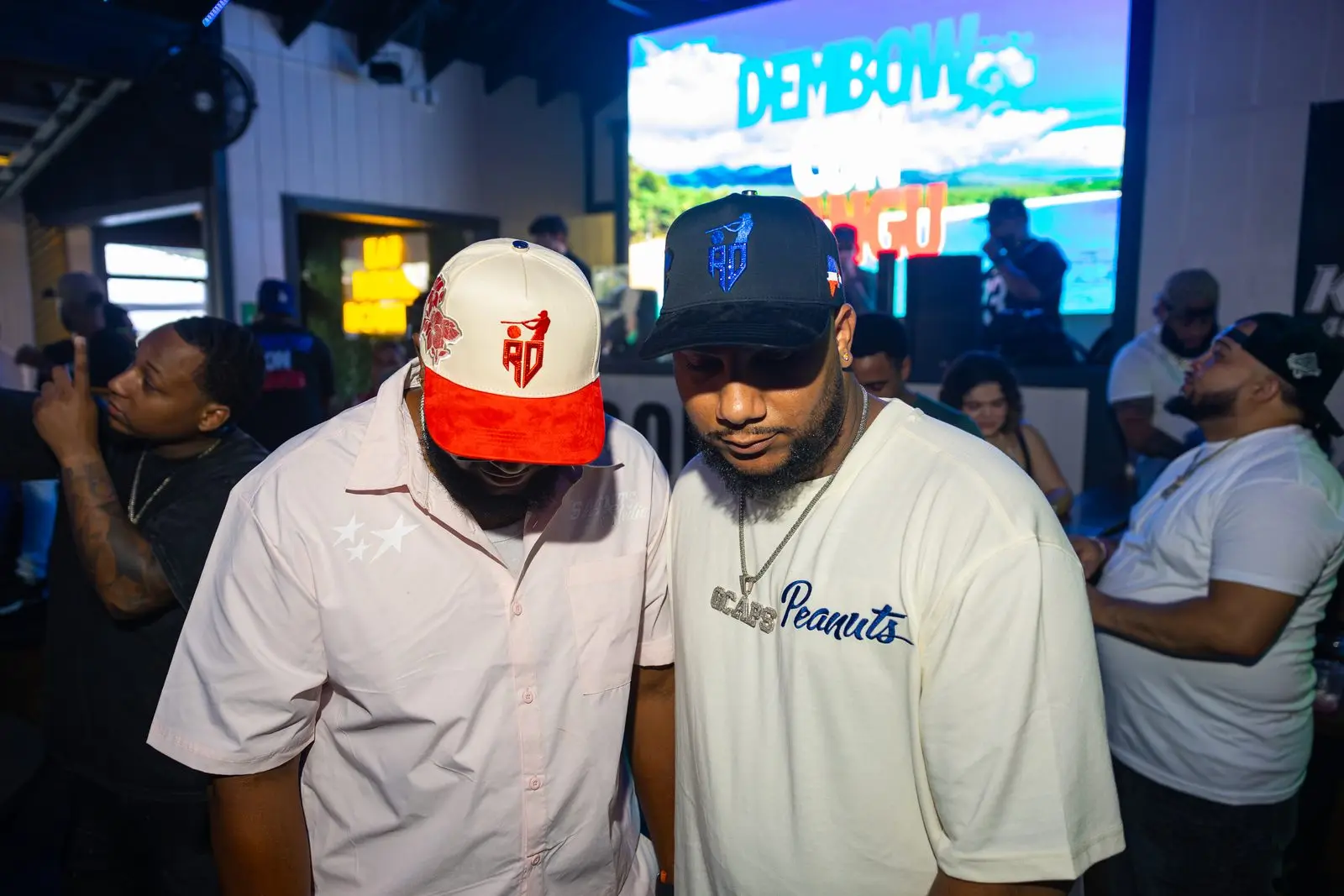 Two men standing close together in a crowded room, the wall-sized screen behind them lit up with the words DEMBOW CON MANGÚ over a green mountainside.