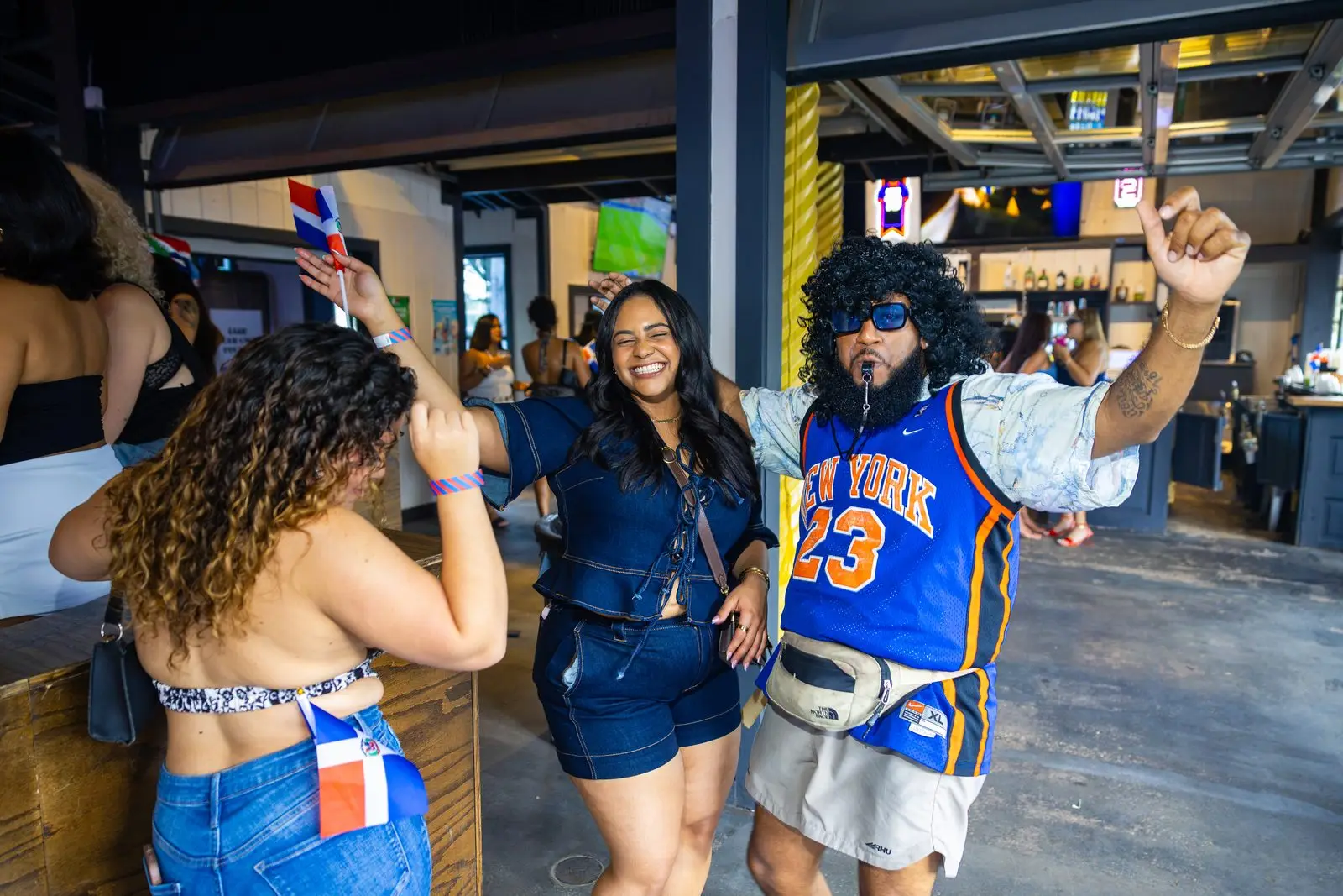 Three people mid-dance with their arms thrown up, one waving a small Dominican flag overhead and another with a flag tucked into a back pocket, an open-sided bar and a row of screens behind them.