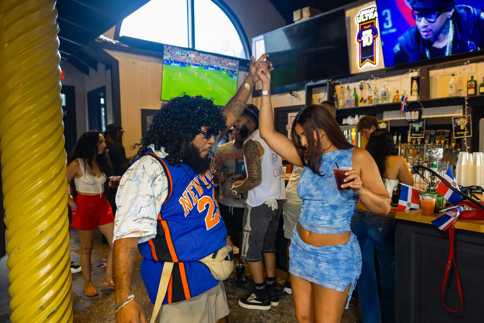 Two people dancing hand in hand in front of the bar, a drink in one hand, bottle shelves and screens behind them and small Dominican flags propped along the bar top.