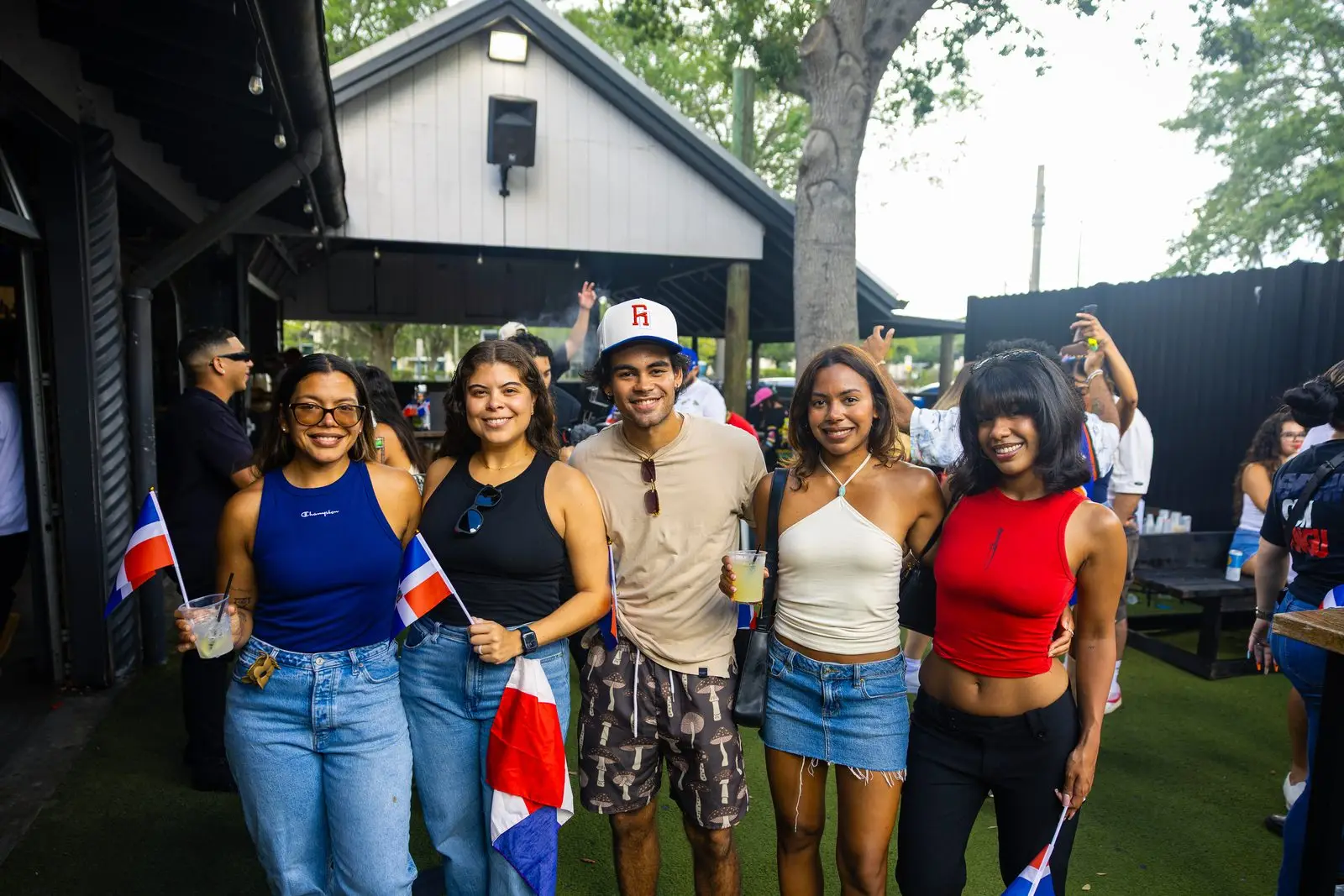 Five friends lined up under an open-sided roof with trees behind them, small Dominican flags in their hands and drinks between them.