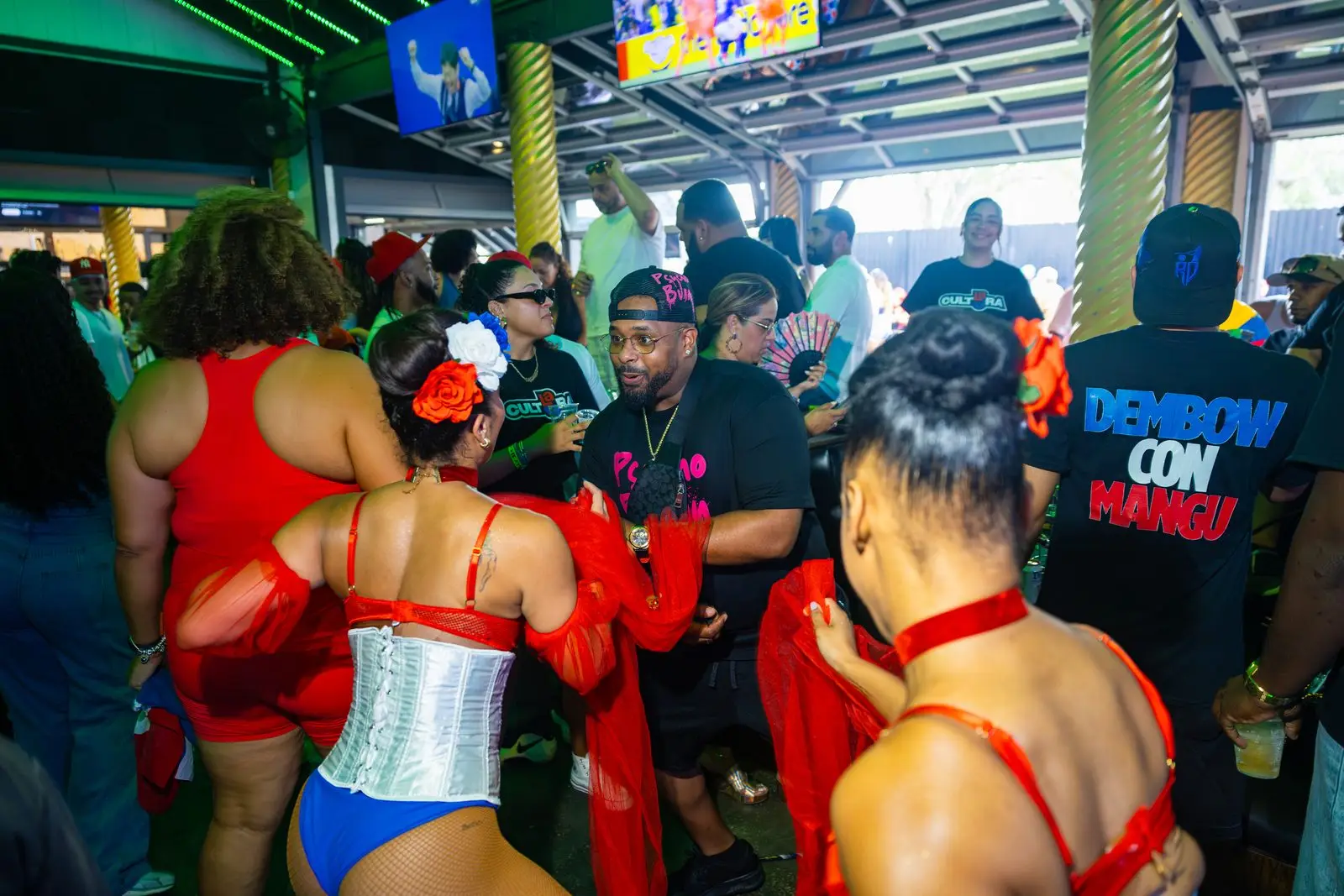 The floor seen from behind the crowd, dancers pressed together front to back, one of them in a Dembow Con Mangú shirt.