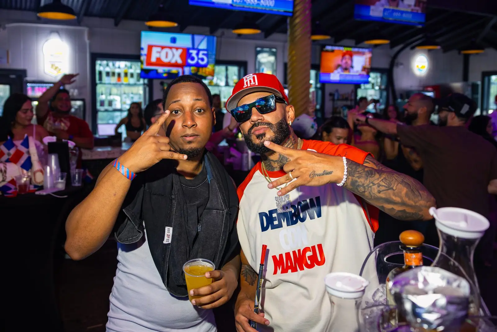 Two men at close range throwing hand signs straight at the camera, one of them in a DEMBOW CON MANGÚ jersey, a bar and a row of television screens behind them.