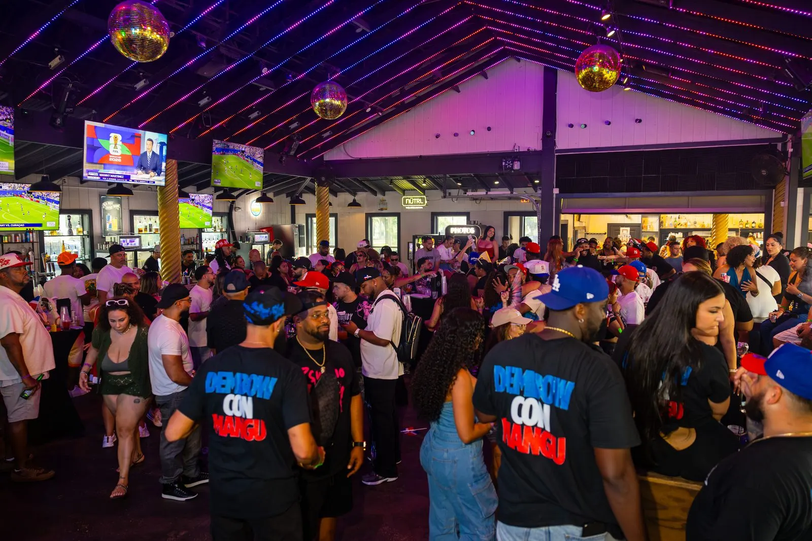 A packed floor under a peaked roof strung with pink and purple lights and mirror balls, several people in Dembow Con Mangú shirts with their backs to the camera.