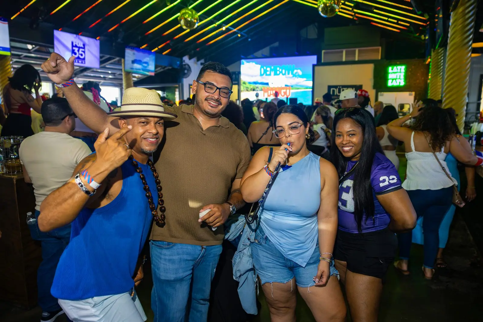 Four friends posing together in a crowded room, one in a straw hat with his fist up, screens and disco balls behind them.