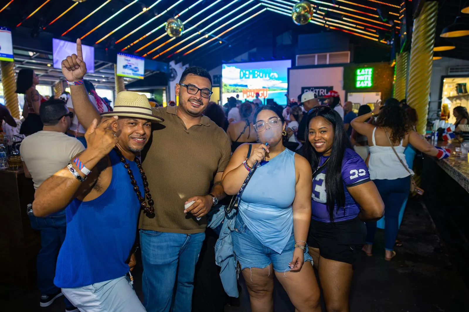 A man in a straw hat with his fist raised mid-celebration, three friends standing with him and a bright screen on the wall behind.