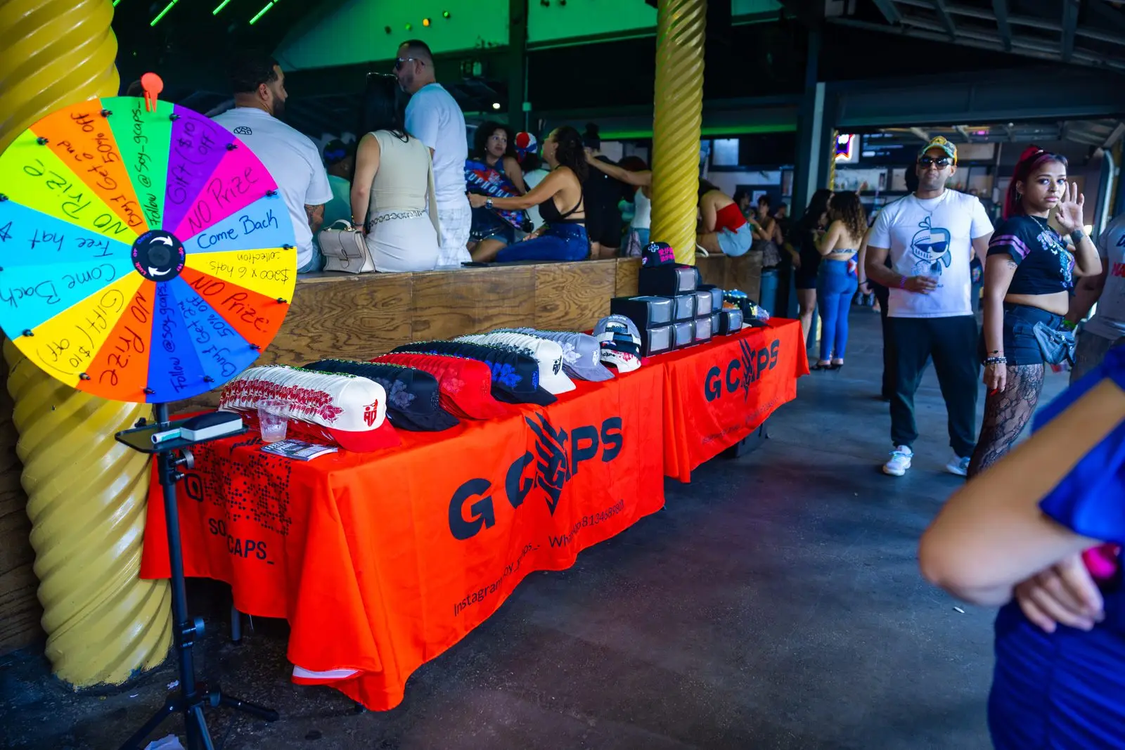 A merchandise table under an open-sided roof, baseball caps laid out across a bright orange cloth, a hand-lettered spin-the-wheel prize game standing at one end of it and people walking past.