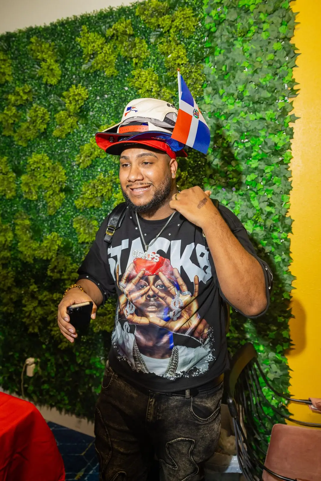 A man smiling straight at the camera in a bucket hat with two small Dominican flags tucked into the brim, against a wall of green foliage.