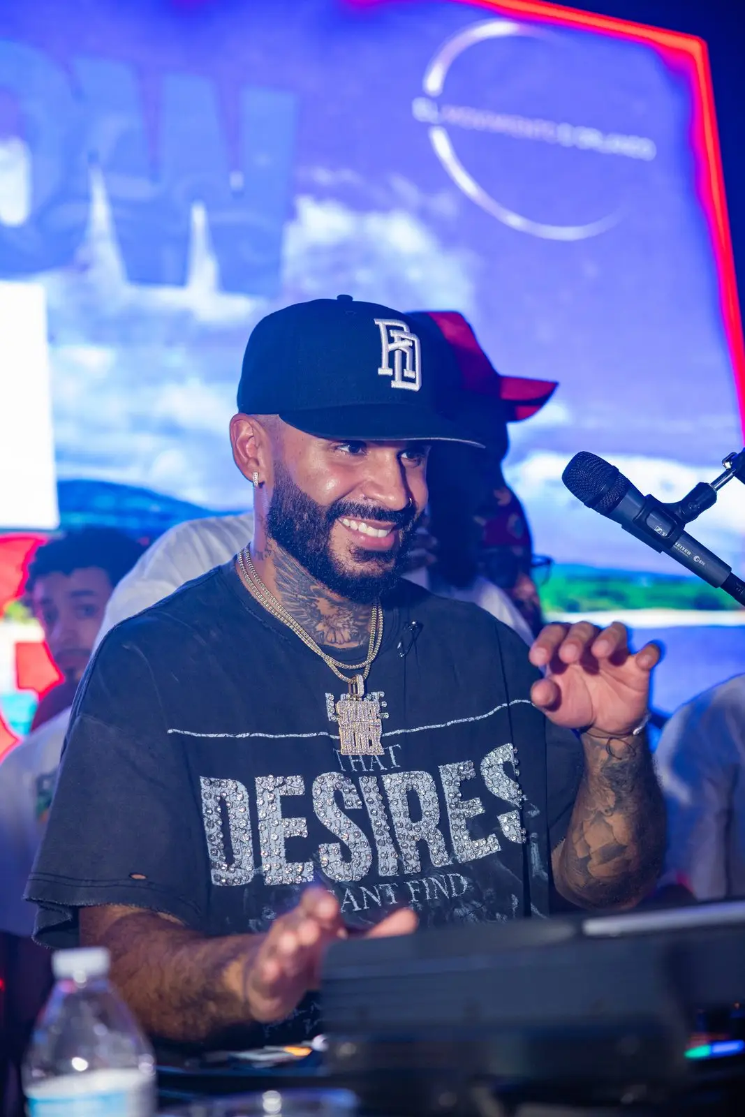 A DJ grinning behind the decks with a microphone at his mouth, the lit Dembow Con Mangú screen filling the wall behind him.