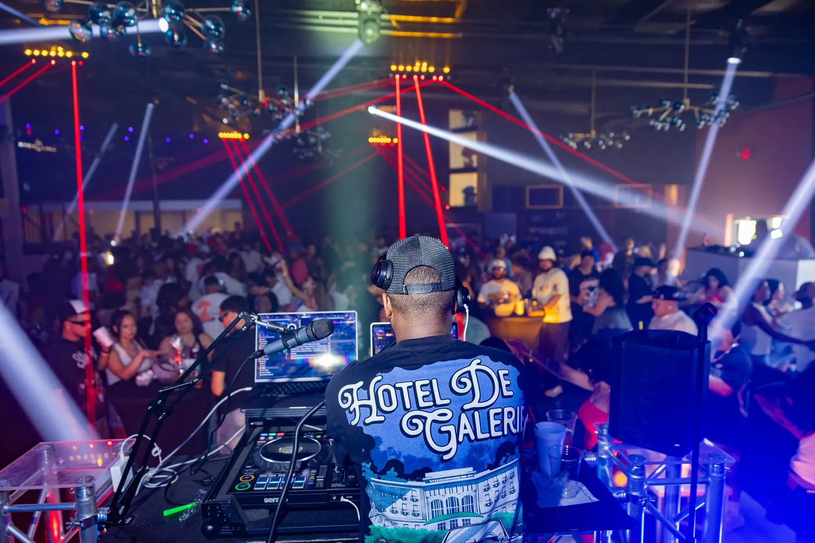A DJ seen from behind at the decks with headphones on, facing a full room, white and red beams crossing above the crowd.