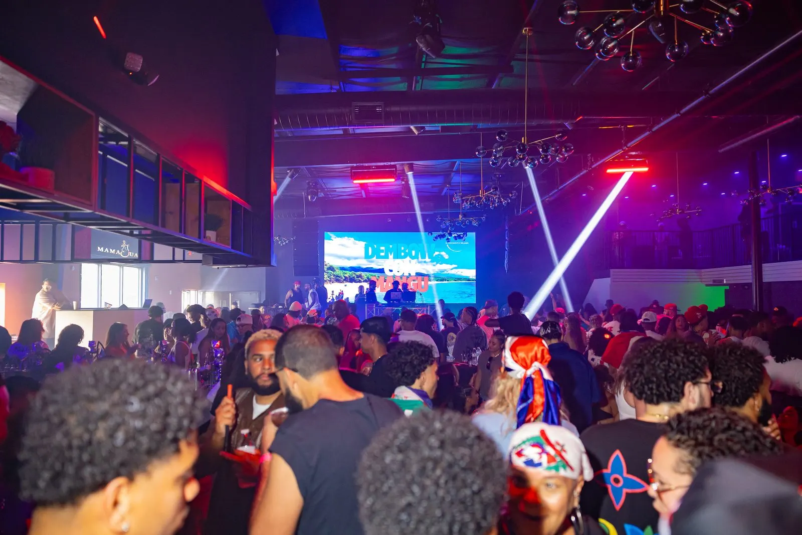 A packed dark room shot from inside the crowd, heads filling the foreground and the DJ booth lit at the far end under a screen reading Dembow Con Mangú.