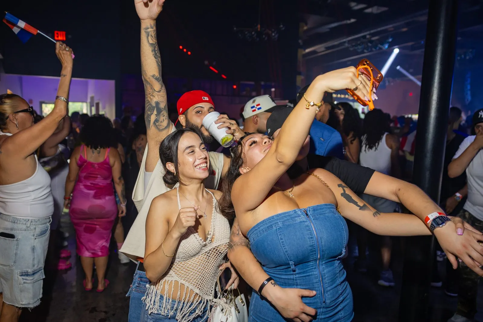 Friends laughing together in a crowded room, one arm raised holding a small Dominican flag, another holding a phone up over the crowd.
