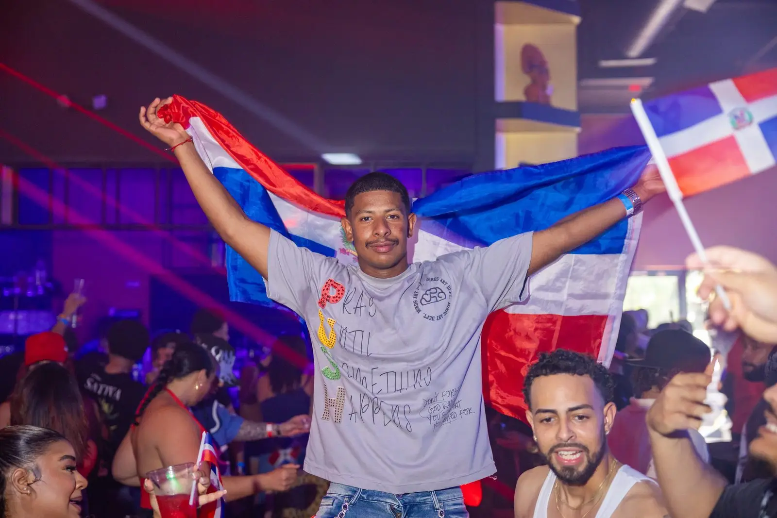 A man holding a Dominican flag open across his outstretched arms, the crowd around him lit purple and red.