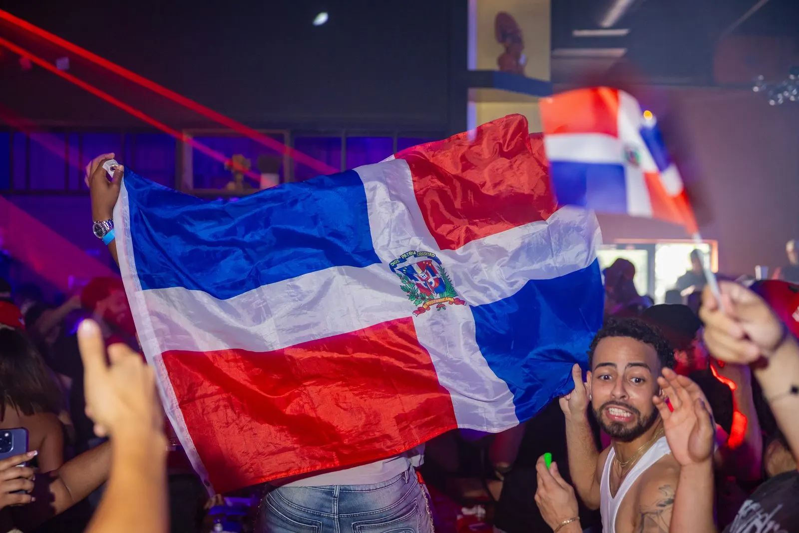 A Dominican flag held open above the crowd at full stretch, a man grinning underneath it and more flags waving behind.